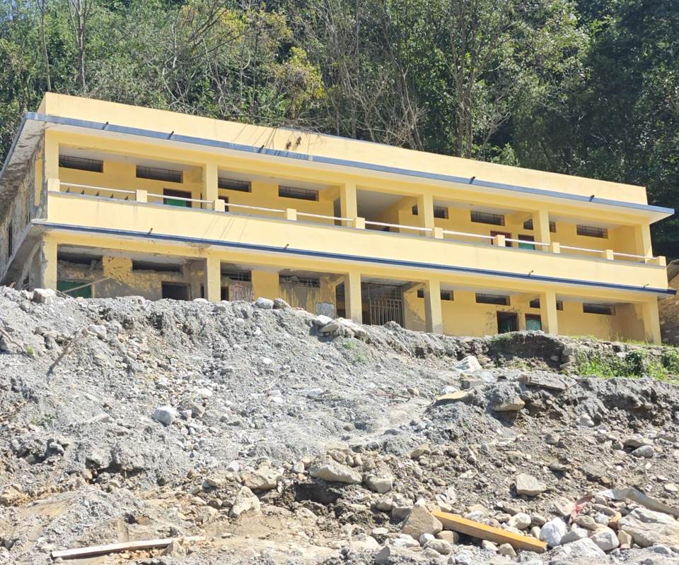 A yellow two-storey school building standing above a slope of flood debris and mud in Rasuwa, Nepal